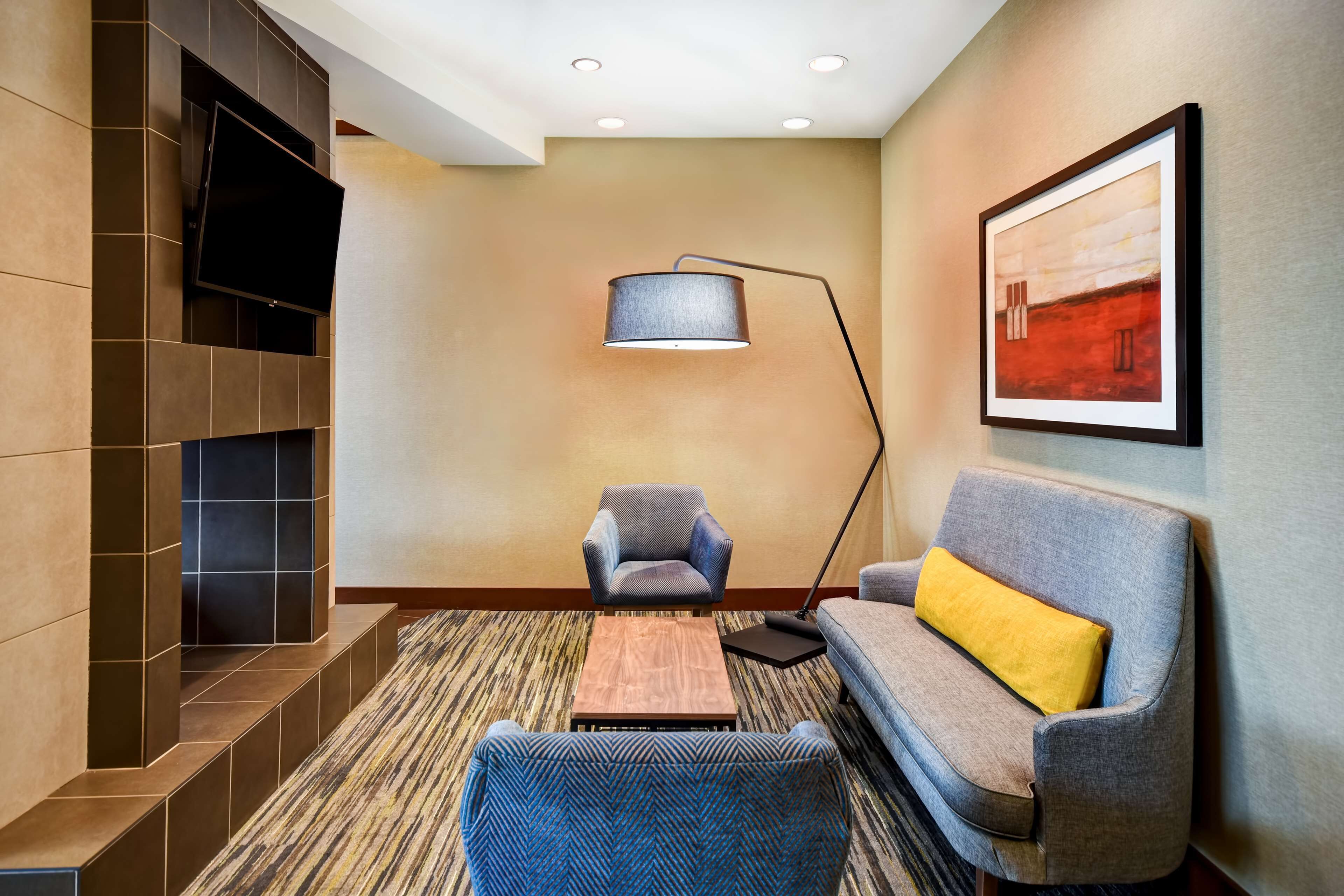 A modern hotel lobby with a flat-screen TV mounted on a wall, a floor lamp, a sofa with a yellow pillow, and a patterned carpet.