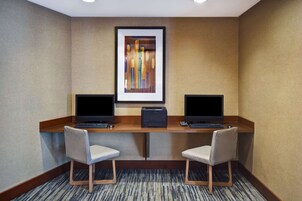 Habitación de hotel con escritorio, dos sillas, dos monitores, una impresora y una obra de arte abstracta enmarcada en la pared.