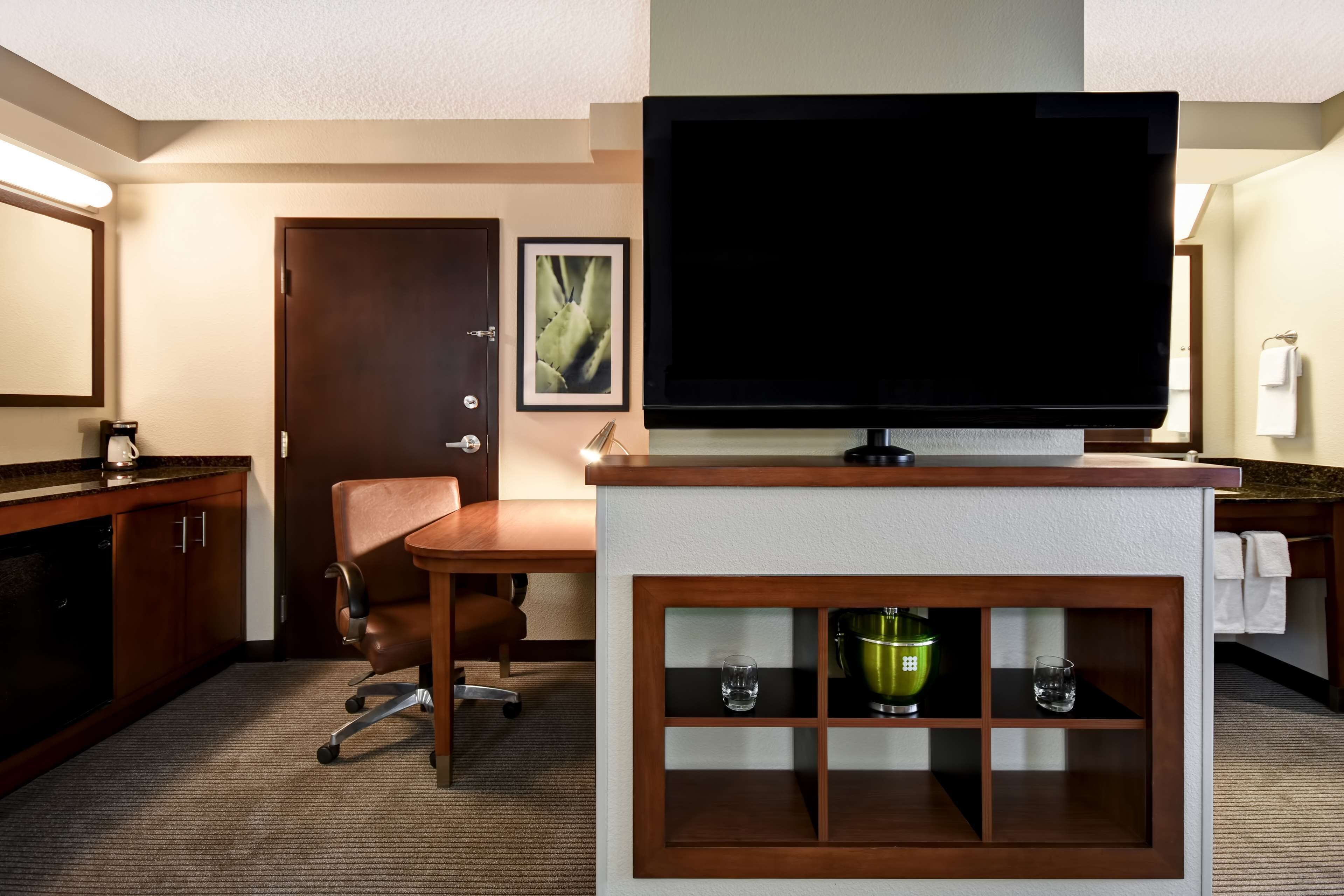 Ein Hotelzimmer mit Fernseher auf einem Standfuß, einem Schreibtisch mit Stuhl, einer Minibar und einer Tür mit einem gerahmten Bild.
