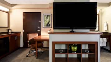 A hotel room with a television on a stand, a desk with a chair, a minibar, and a door with a framed picture.