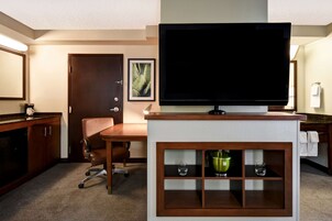 A hotel room with a television on a stand, a desk with a chair, a minibar, and a door with a framed picture.