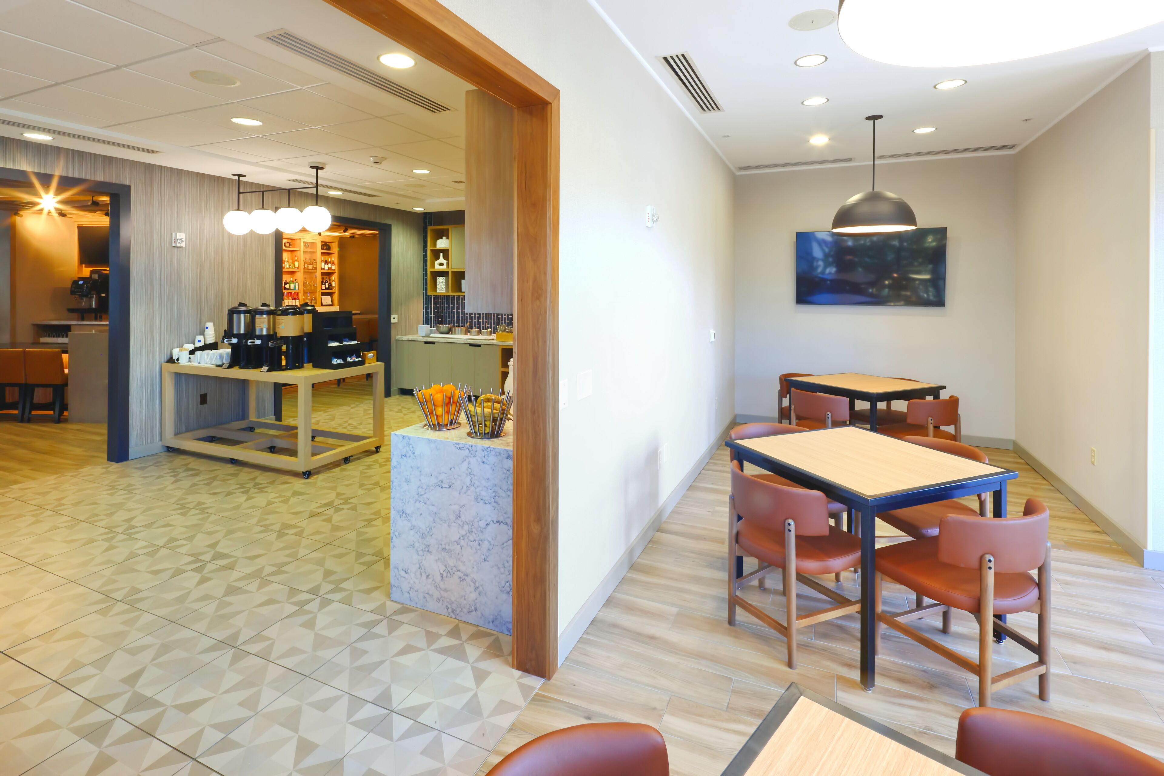 A modern hotel lobby with a dining area, a coffee station, and a kitchen in the background.