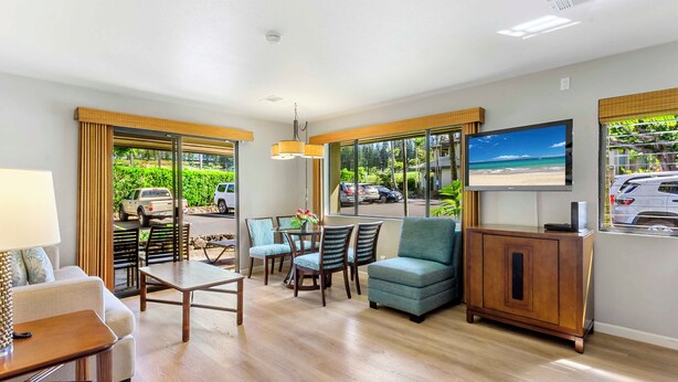 A hotel room with a sofa, a dining table with chairs, a TV mounted on the wall, and large windows offering a view of the outside. - The Gardens at West Maui (Lahaina)
