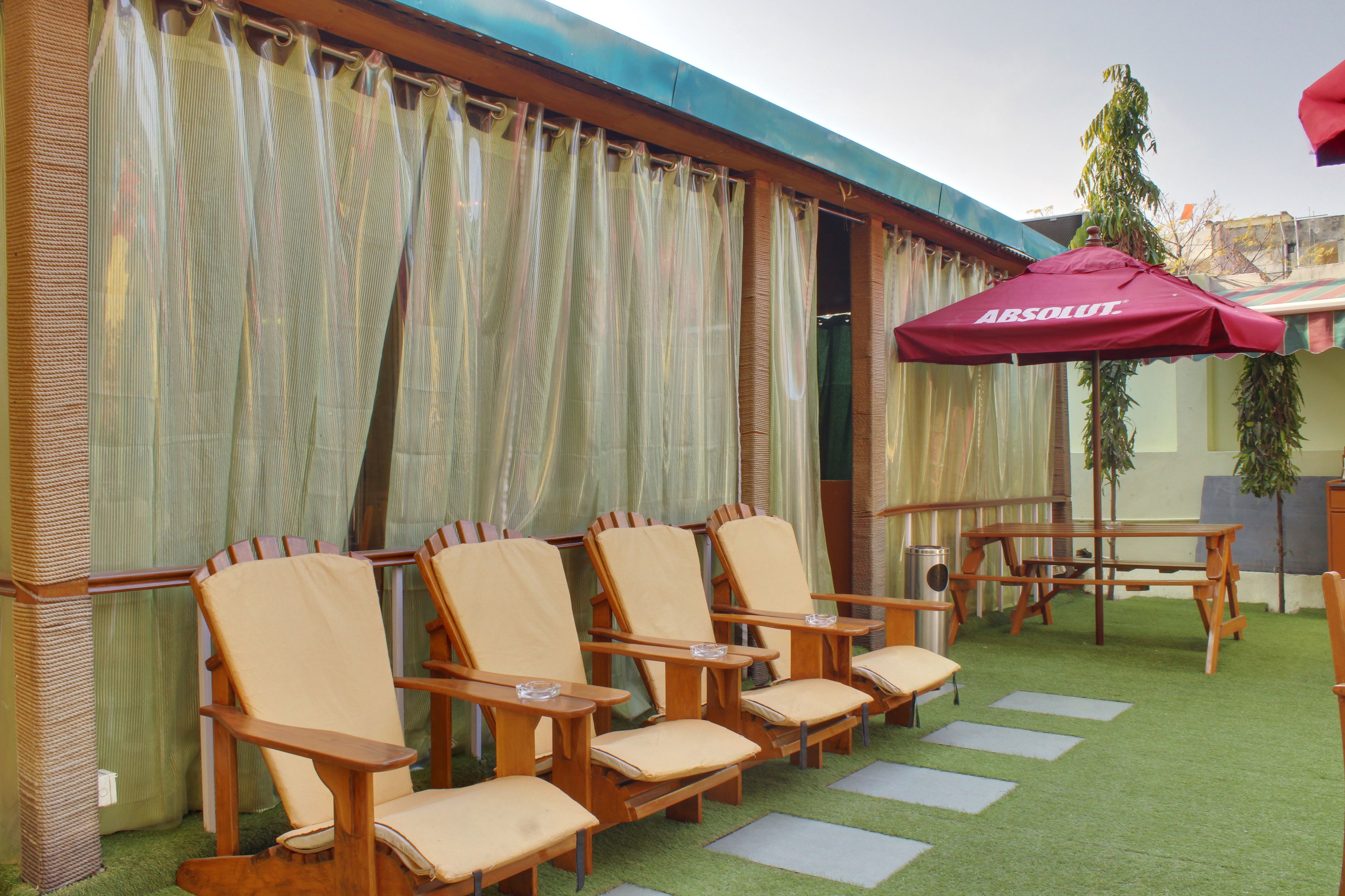 A seating area with wooden chairs and a table, a red umbrella, and a view of a building through sheer curtains.