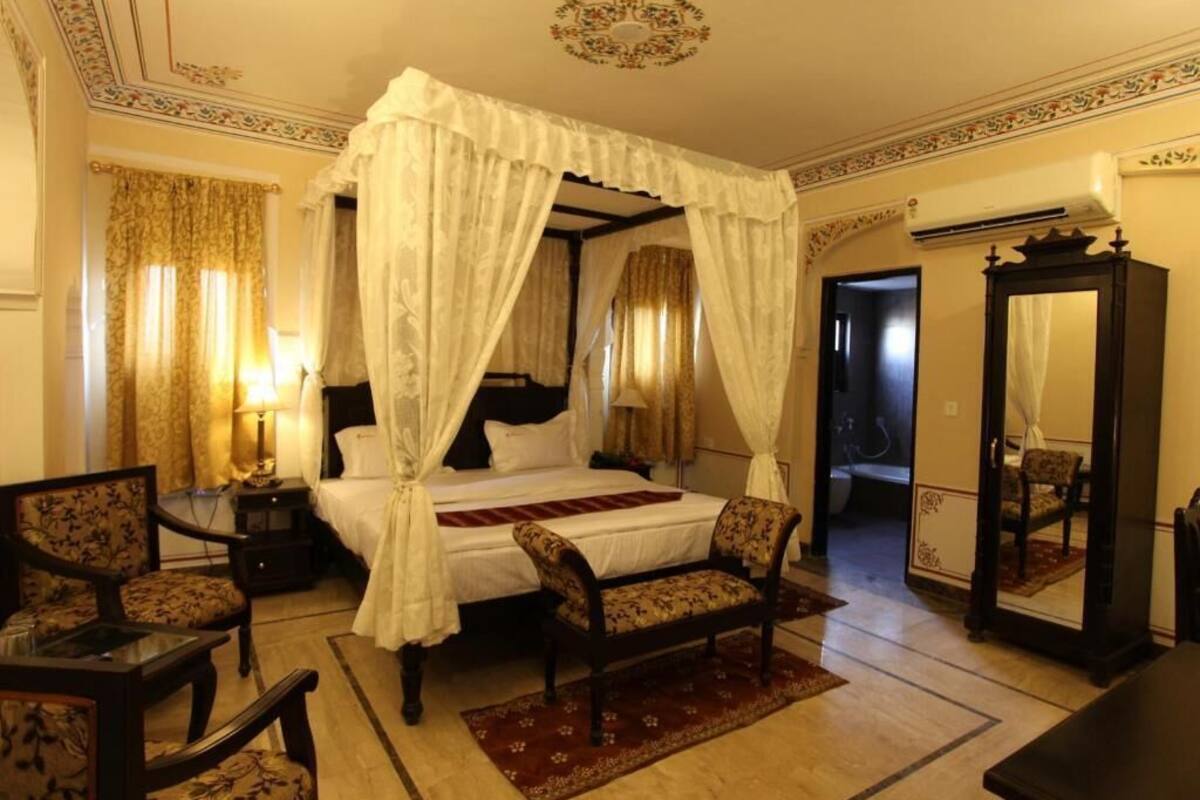A four-poster canopy bed with a red throw, two upholstered chairs, a mirror, and a nightstand with a lamp.