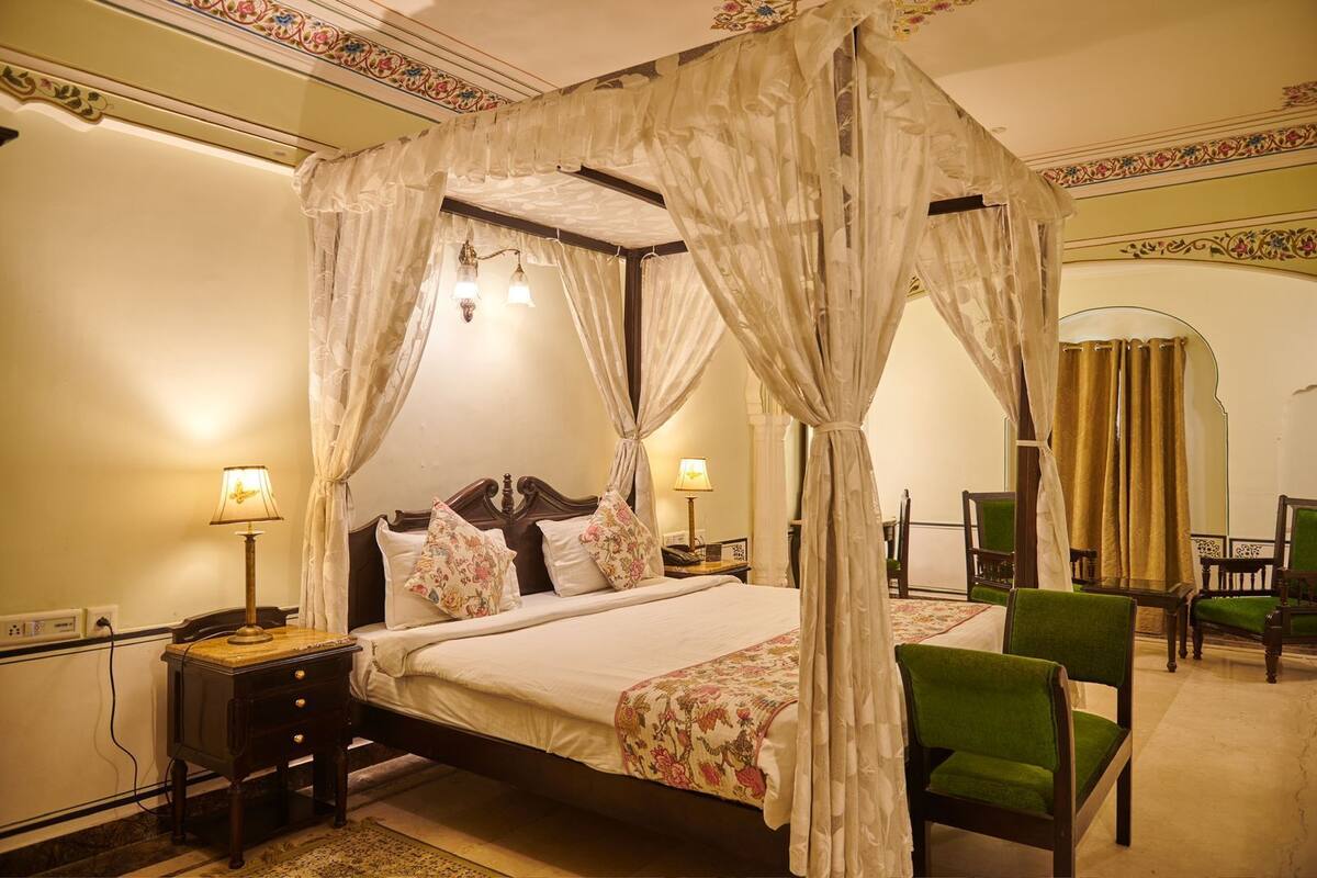 A four-poster canopy bed with floral bedding, a nightstand with a lamp, and a green armchair.