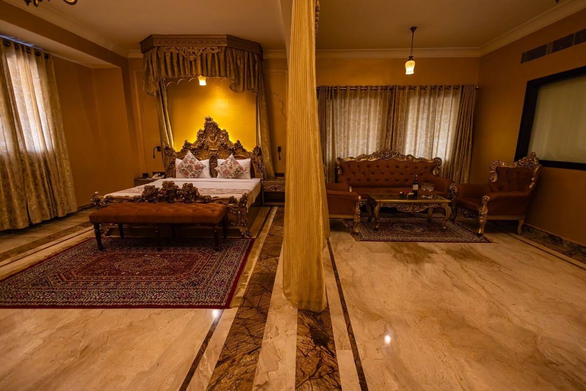 A luxurious bedroom with a large bed, ornate headboard, a sitting area with a sofa and armchairs, and a large window with curtains.