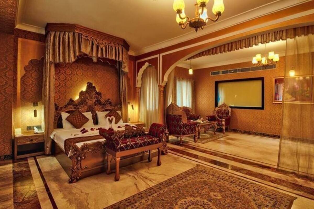 A luxurious bedroom with a large bed, ornate headboard, a sitting area with a sofa and armchairs, and a large window with curtains.
