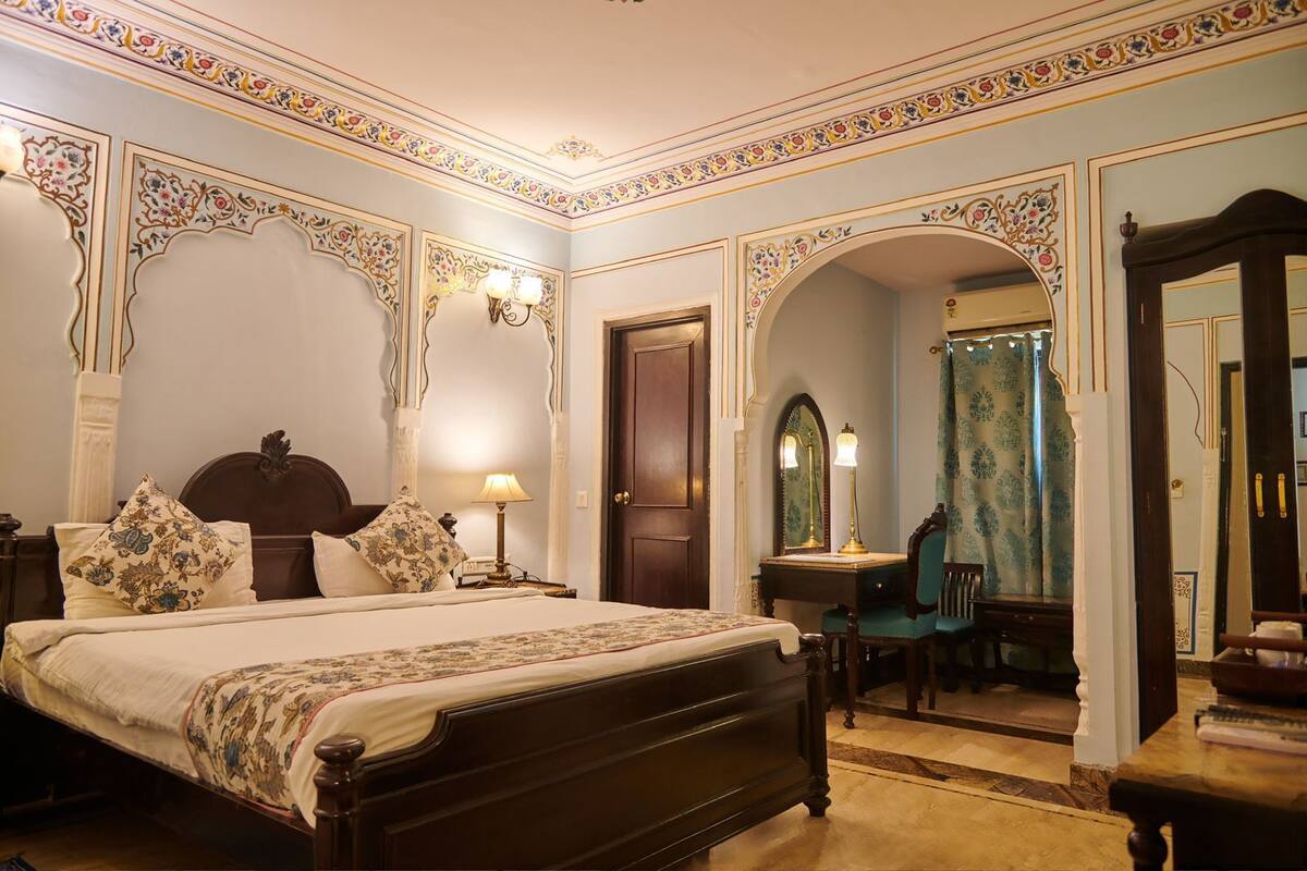 A traditional bedroom with a large bed, ornate wall decorations, a desk with a chair, and a mirror.