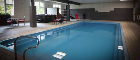An indoor swimming pool with a tiled floor, a pool ladder, and a seating area with chairs and tables.