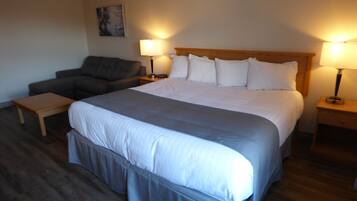 A hotel room with a large bed, two bedside tables with lamps, a grey sofa, and a small wooden table.