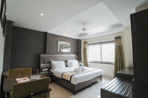 A hotel room with a bed, a desk, a chair, a window with curtains, and a wall-mounted mirror.