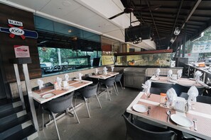 L’intérieur d’un restaurant, avec des tables dressées pour le service, une vue à travers la fenêtre et une enseigne indiquant le nom de l’établissement.