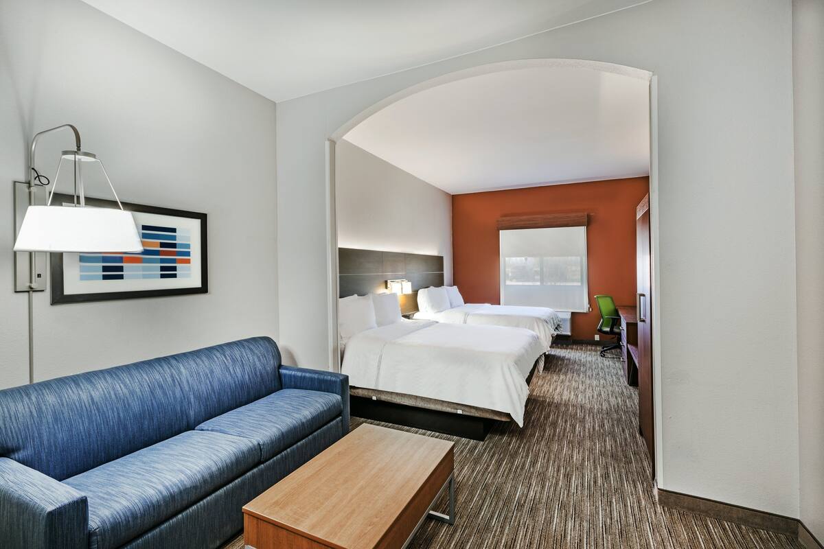 A hotel room with a blue sofa, a wooden coffee table, a bed with white bedding, a desk with a chair, and a framed picture on the wall.