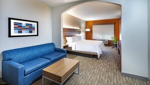 Ein Hotelzimmer mit einem blauen Sofa, einem Holztisch, einem Bett mit weißen Leinentüchern, einem grünen Sessel und einem gerahmten abstrakten Kunstwerk an der Wand.