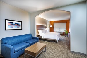 Une chambre d’hôtel avec un canapé bleu, une table basse en bois, un lit avec du linge de lit blanc, une chaise verte et une œuvre d’art abstraite encadrée accrochée au mur.
