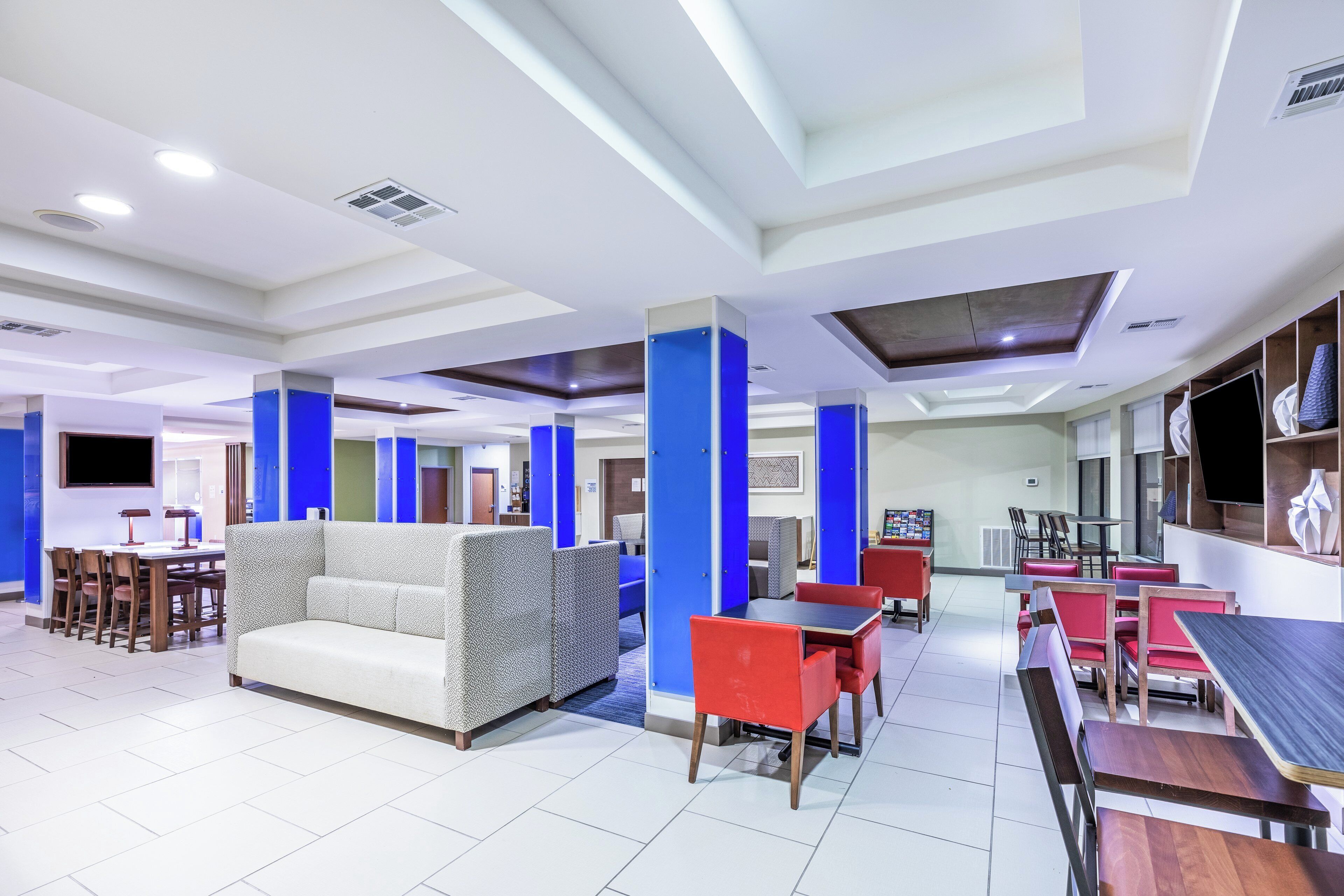 A modern hotel lobby with a central seating area, dining tables, and a flat-screen TV mounted on the wall.