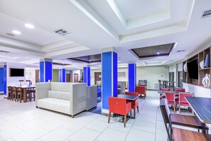 Un moderno hall de hotel con un área de estar central, mesas de comedor y un televisor de pantalla plana en la pared.