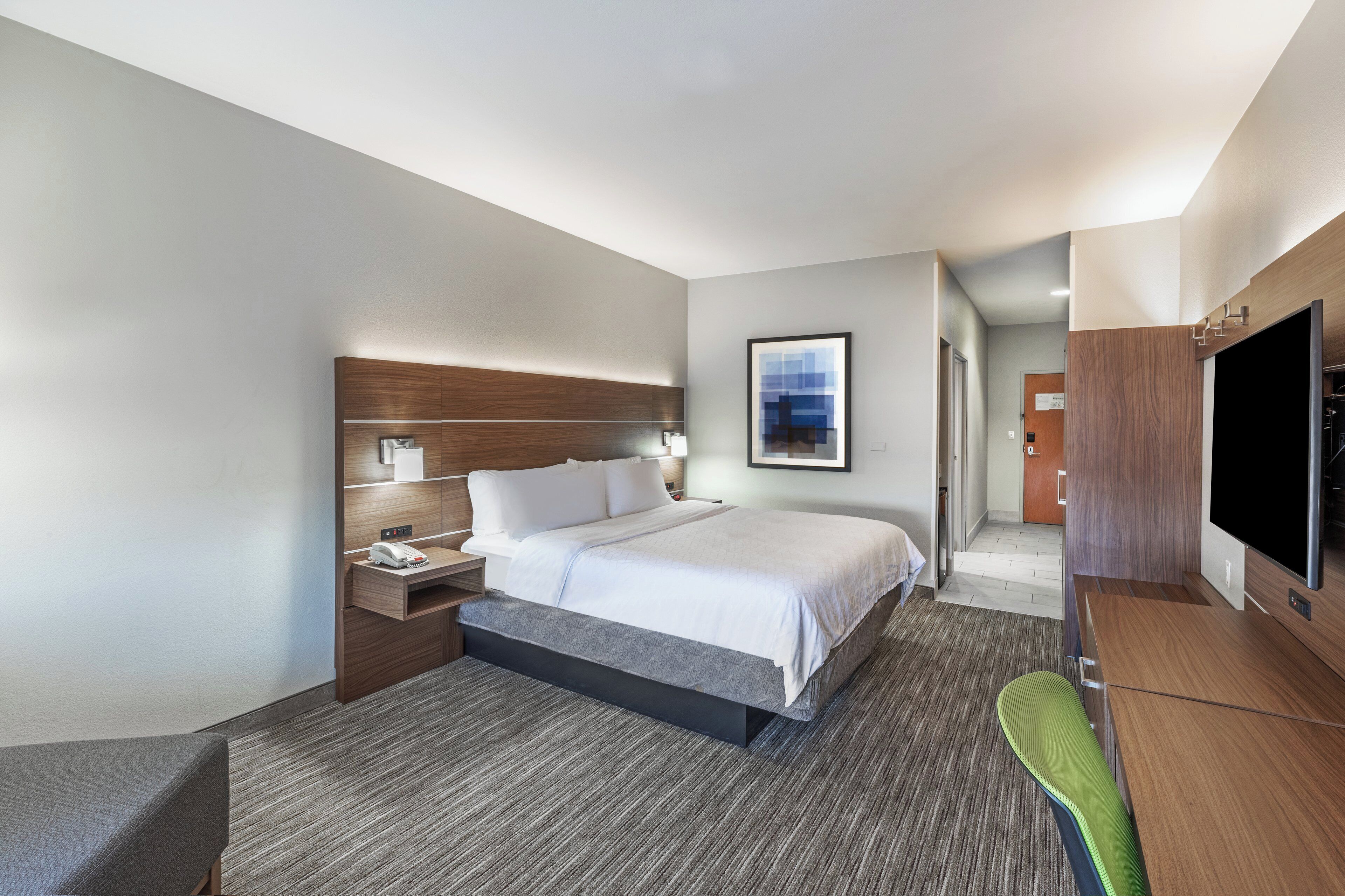 A hotel room with a bed, bedside table, lamp, artwork, television, and a green chair.