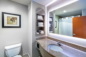 Une salle de bain avec des toilettes blanches, un lavabo en granit, un grand miroir et des étagères où sont pliées des serviettes.