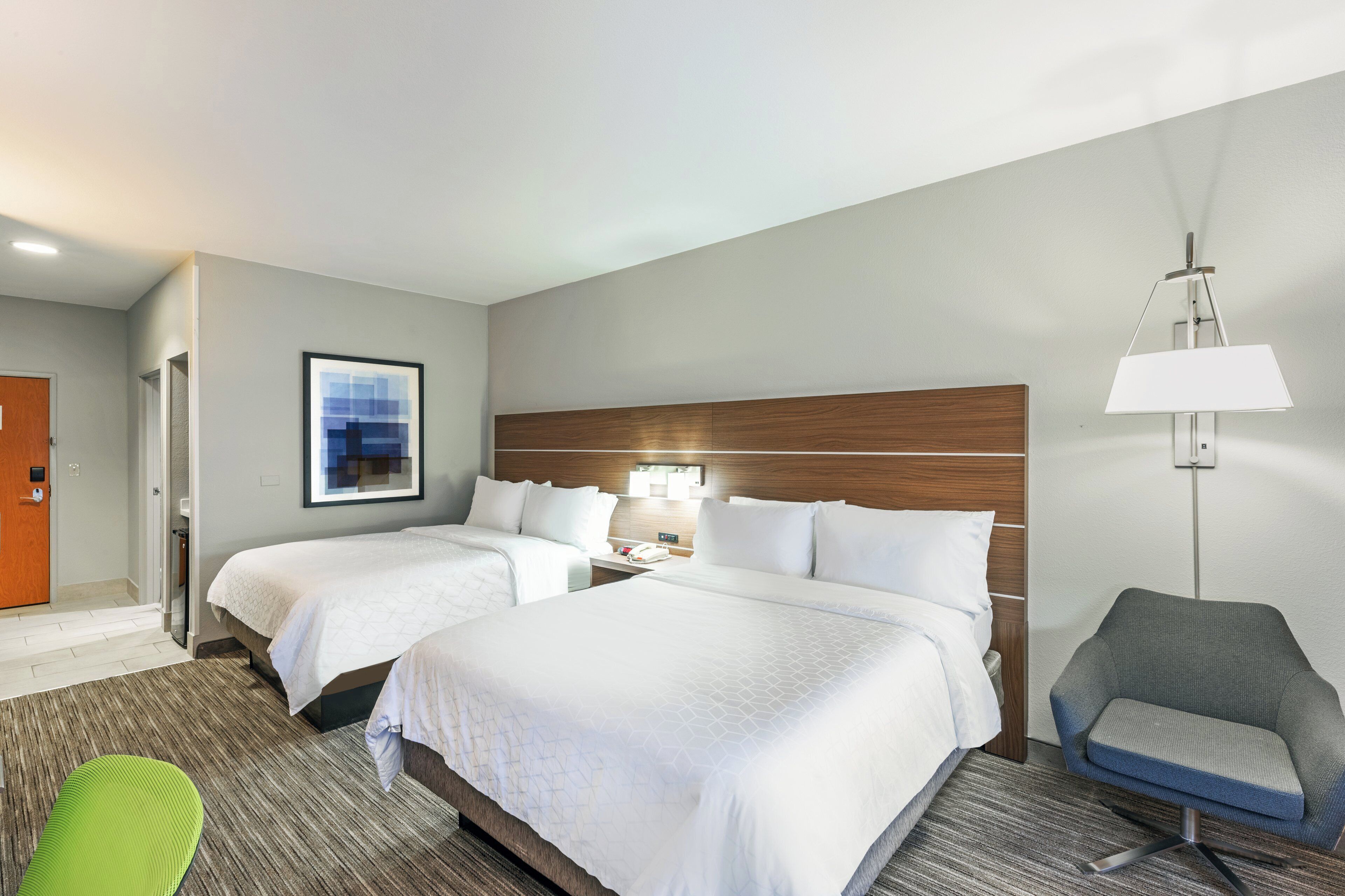 A hotel room with two beds, a desk, a chair, and a lamp.