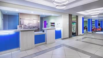 A hotel reception area with a modern design, including a reception desk, a self-service kiosk, and a seating area with tables and chairs.