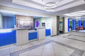 Un espace d’accueil d’hôtel au design moderne, comprenant un comptoir d’accueil, un kiosque en libre-service et un coin salon avec des tables et des chaises.