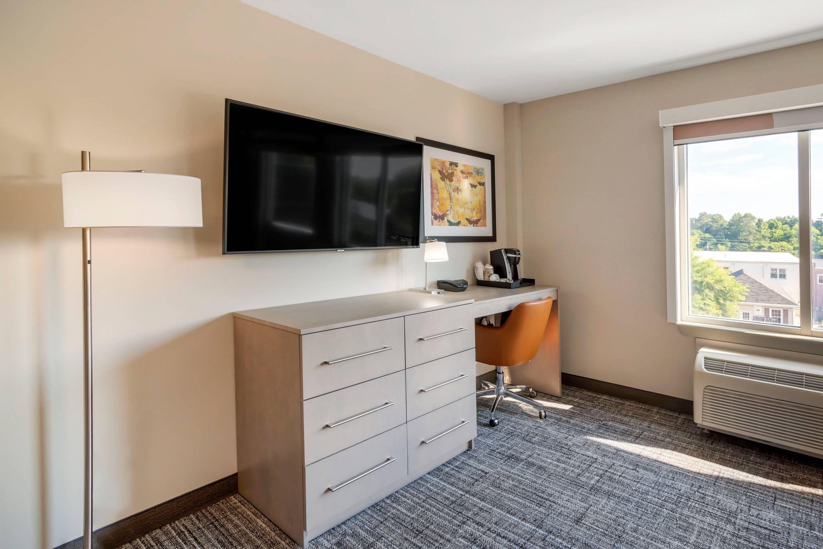 A hotel room with a flat-screen TV mounted above a desk, a large window with a view of buildings, and a carpeted floor.