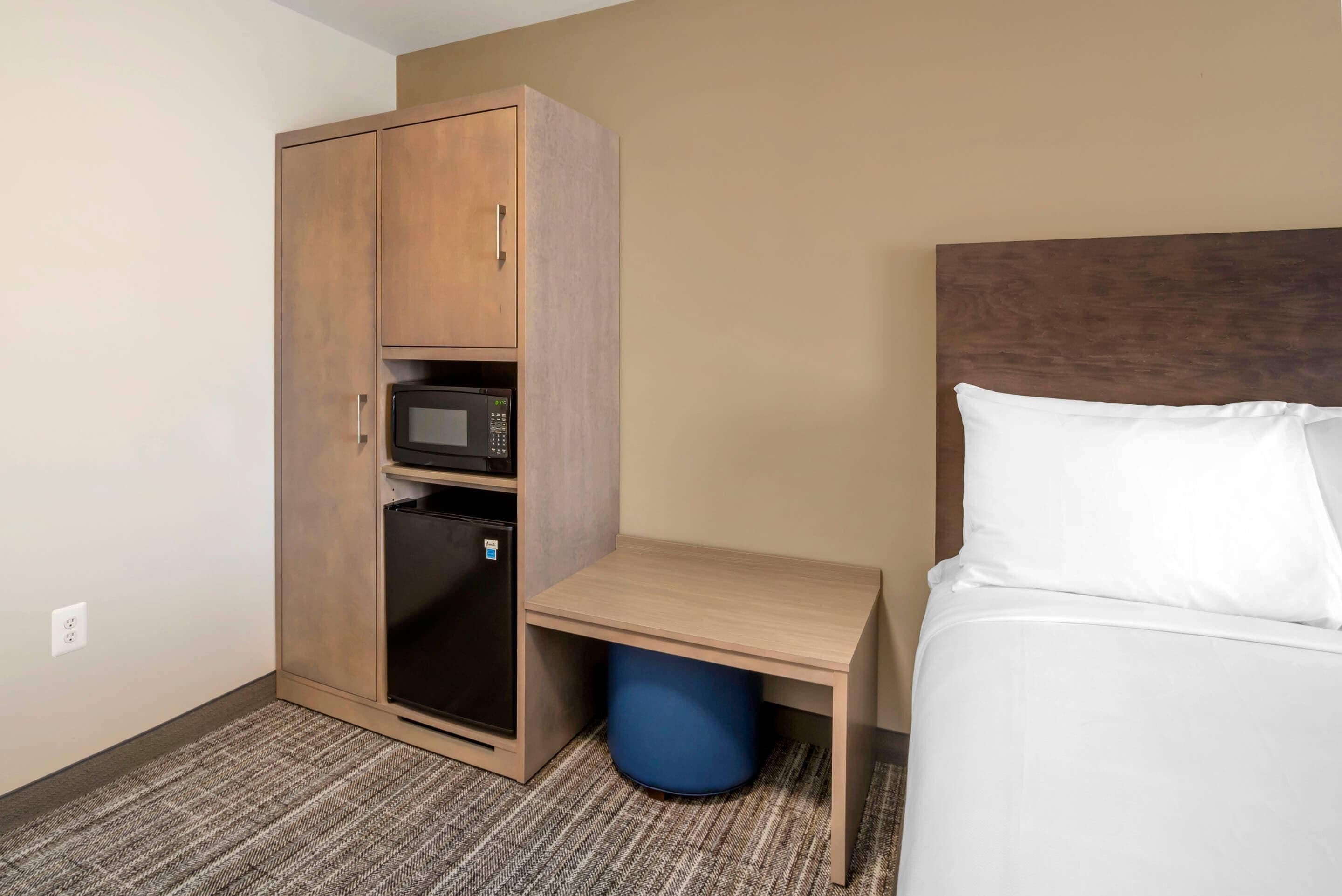 Une chambre d’hôtel avec un lit, une armoire en bois équipée d’un micro-ondes, une petite table et une poubelle bleue.