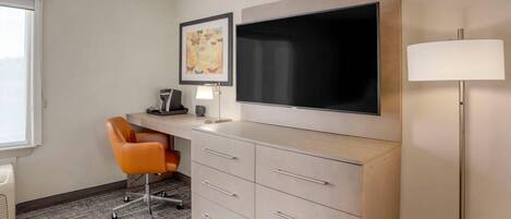 A hotel room with a desk, chair, flat-screen TV, and a lamp.