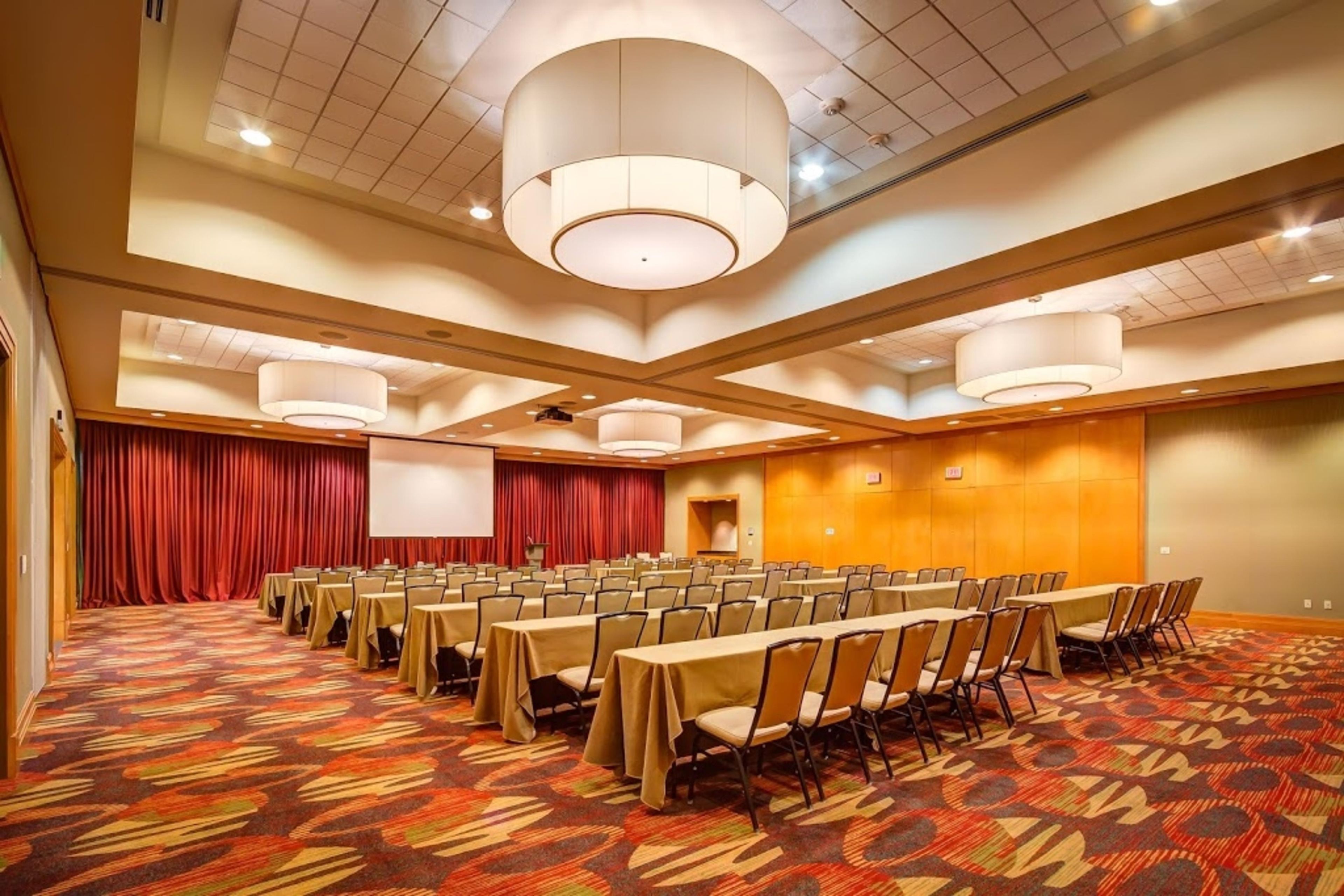 Een congreszaal met rijen tafels en stoelen, een podium met gordijnen, en een groot scherm.