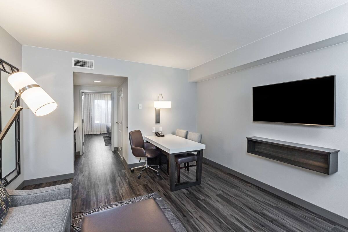 A modern hotel room with a wooden floor, a dining area with a table and chairs, a flat-screen TV mounted on the wall, and a hallway leading to another room.