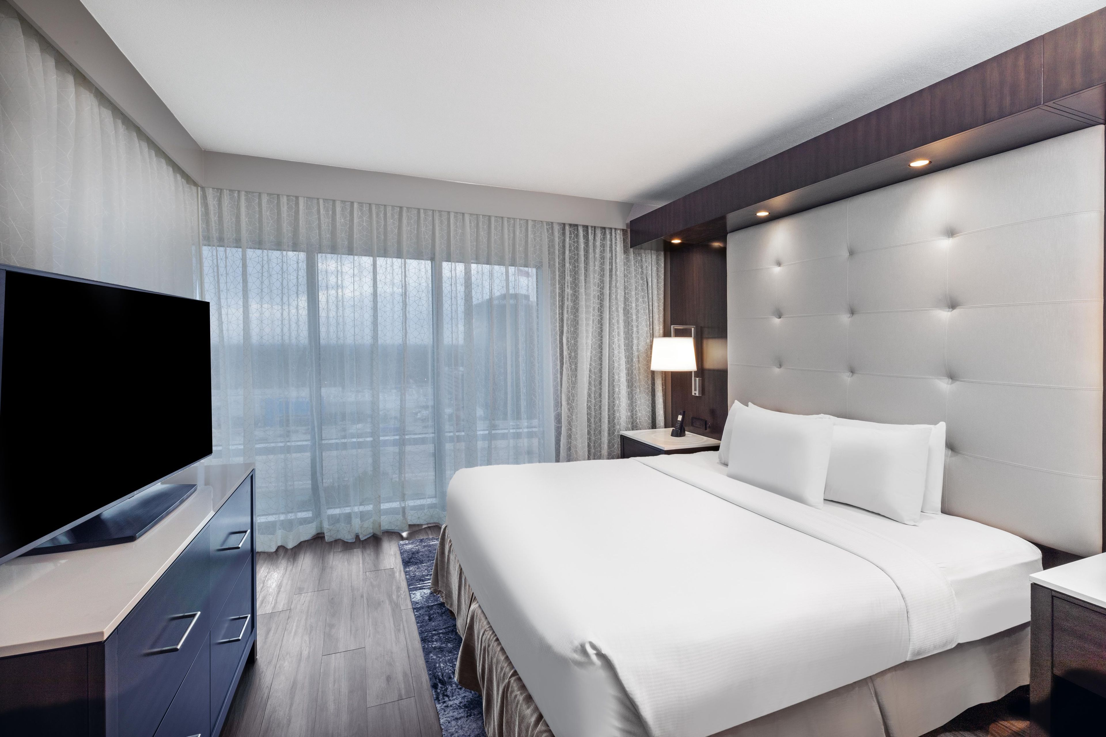Habitación de hotel con una cama grande, un televisor de pantalla plana, una mesita de noche con lámpara y una ventana con cortinas translúcidas.