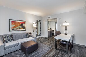 A modern hotel room with a grey sofa, a small ottoman, a dining table with chairs, a kitchen area, and a large mirror.