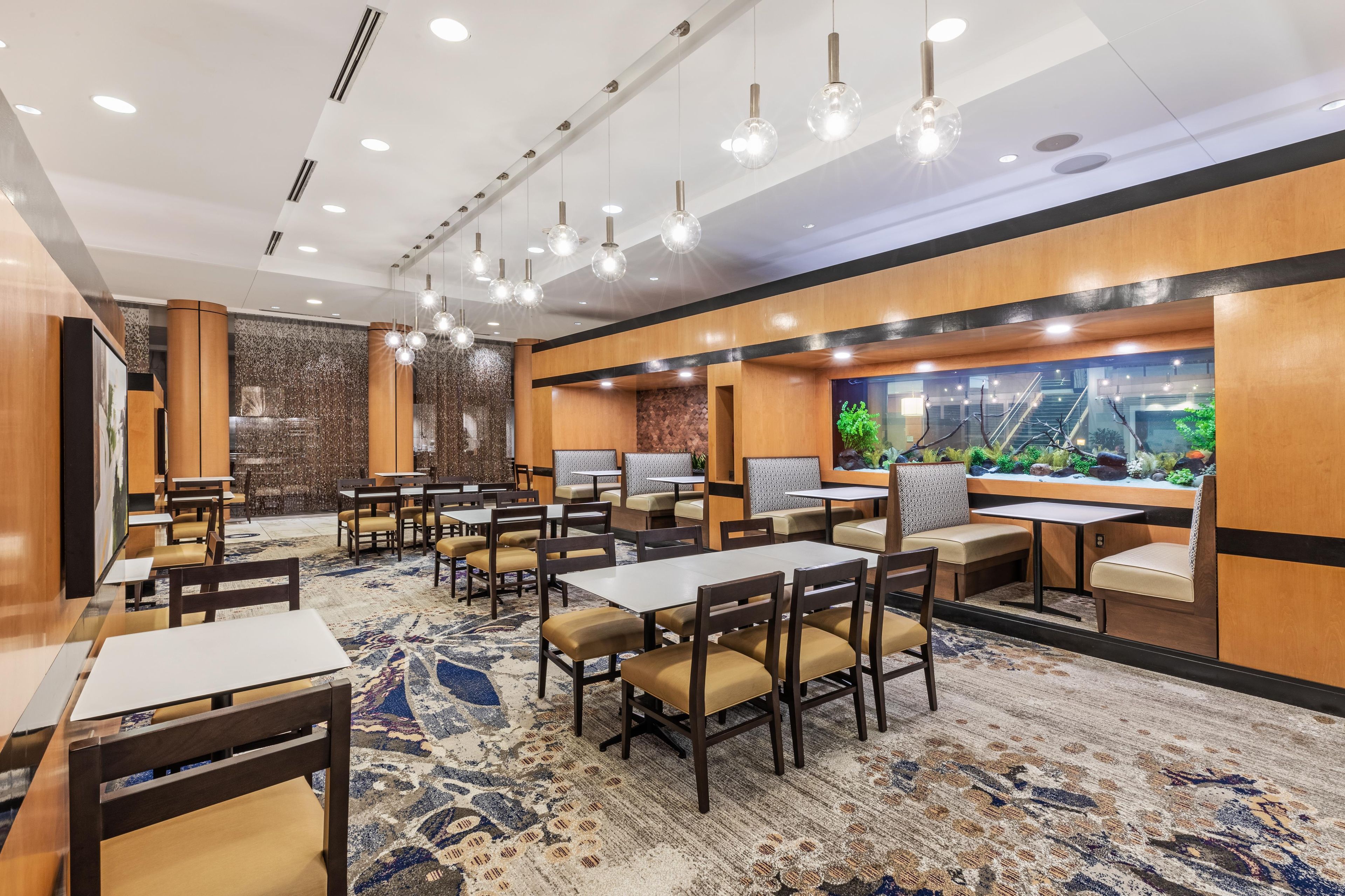 A hotel dining area with multiple tables and booths, a patterned carpet, and a large aquarium in the background.