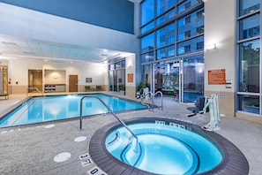An indoor swimming pool with a hot tub, seating area, and a view of a building with large windows.