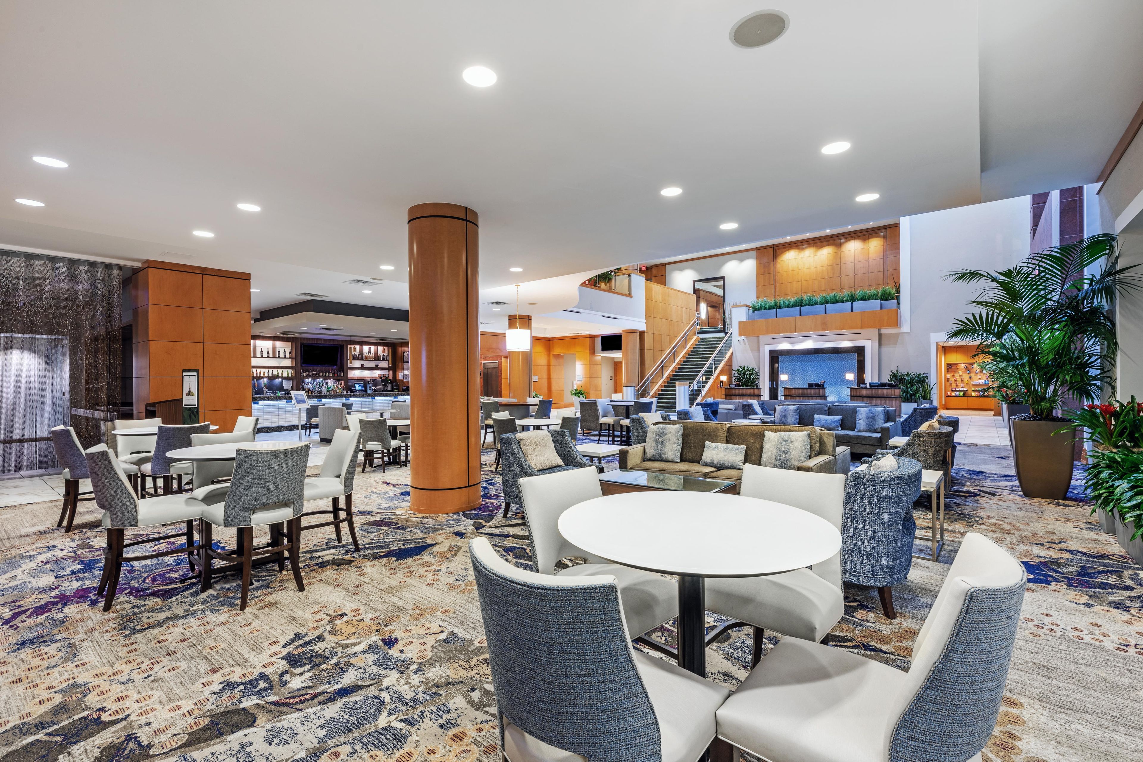 Un hall de hotel con una zona de bar, disposición de asientos y una escalera que lleva a un nivel superior.
