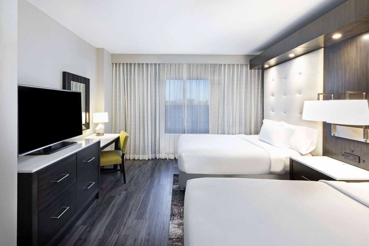 A modern hotel room with a flat-screen TV, a desk with a chair, two beds, a large window with curtains, and a headboard with built-in lighting.