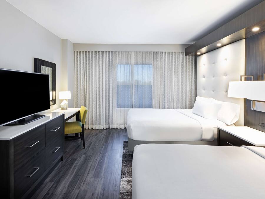 A modern hotel room with a flat-screen TV, a desk with a chair, two beds, a large window with curtains, and a headboard with built-in lighting.