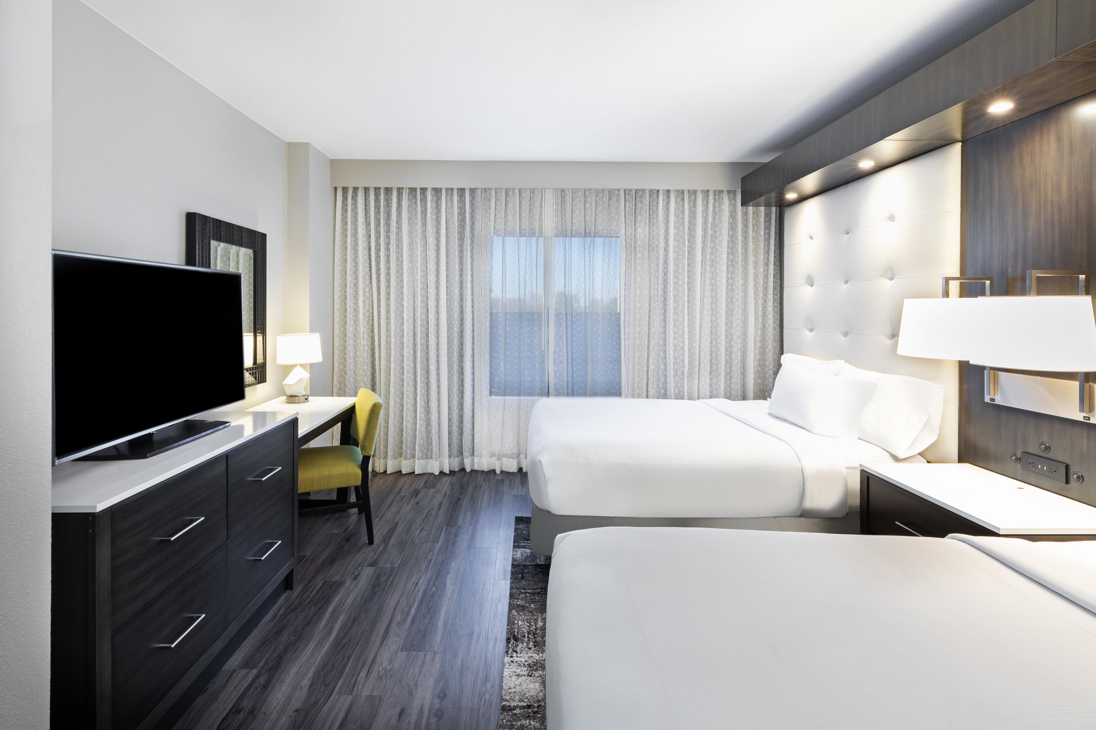 A hotel room with a flat-screen TV, a desk with a lamp, two beds, and a large window with curtains.