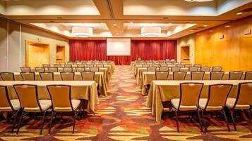 Un salón de conferencias con filas de mesas y sillas, un escenario con cortina roja y una señal de salida.
