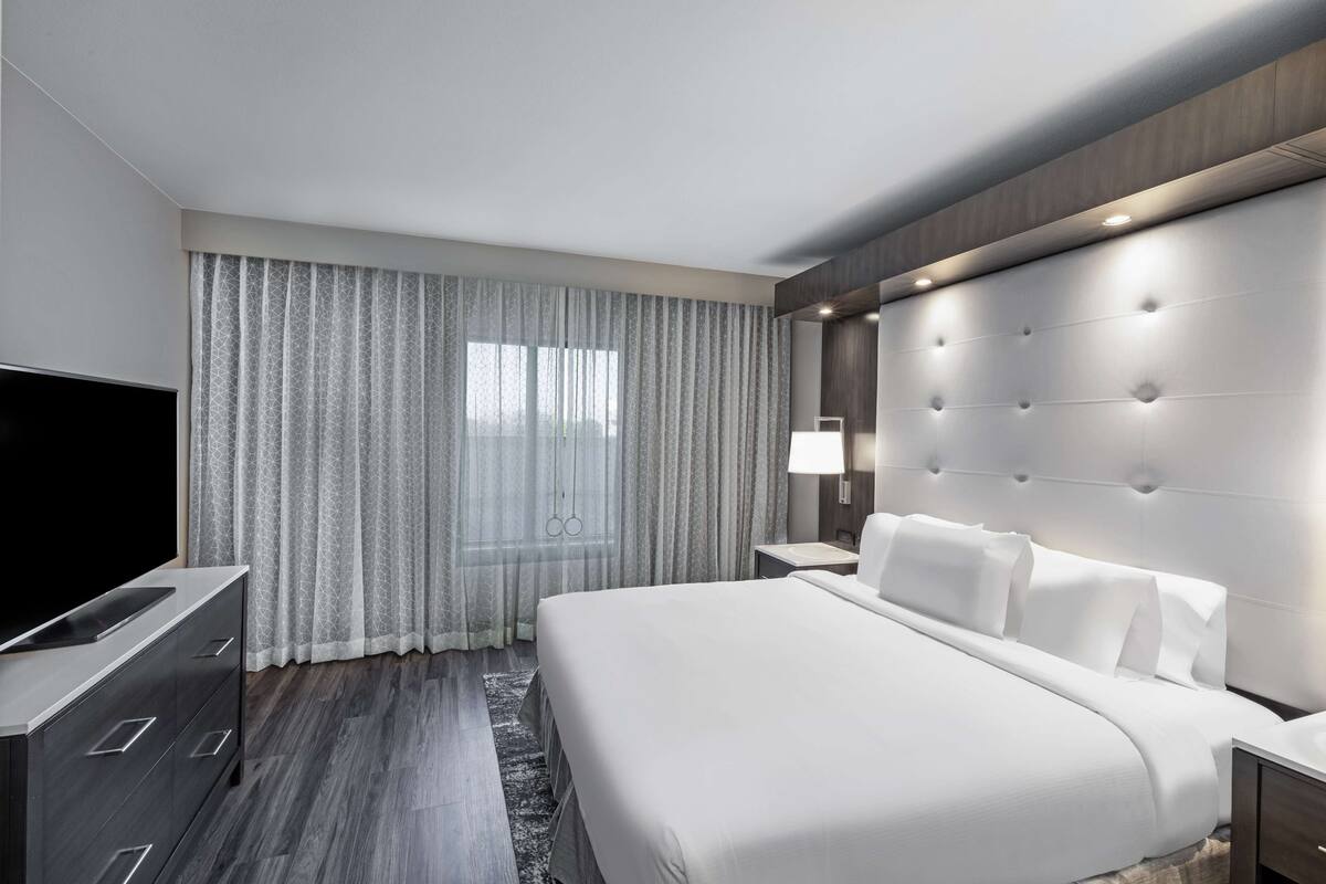 A modern hotel room with a large bed, a flat-screen TV, a bedside table, and a window with curtains.
