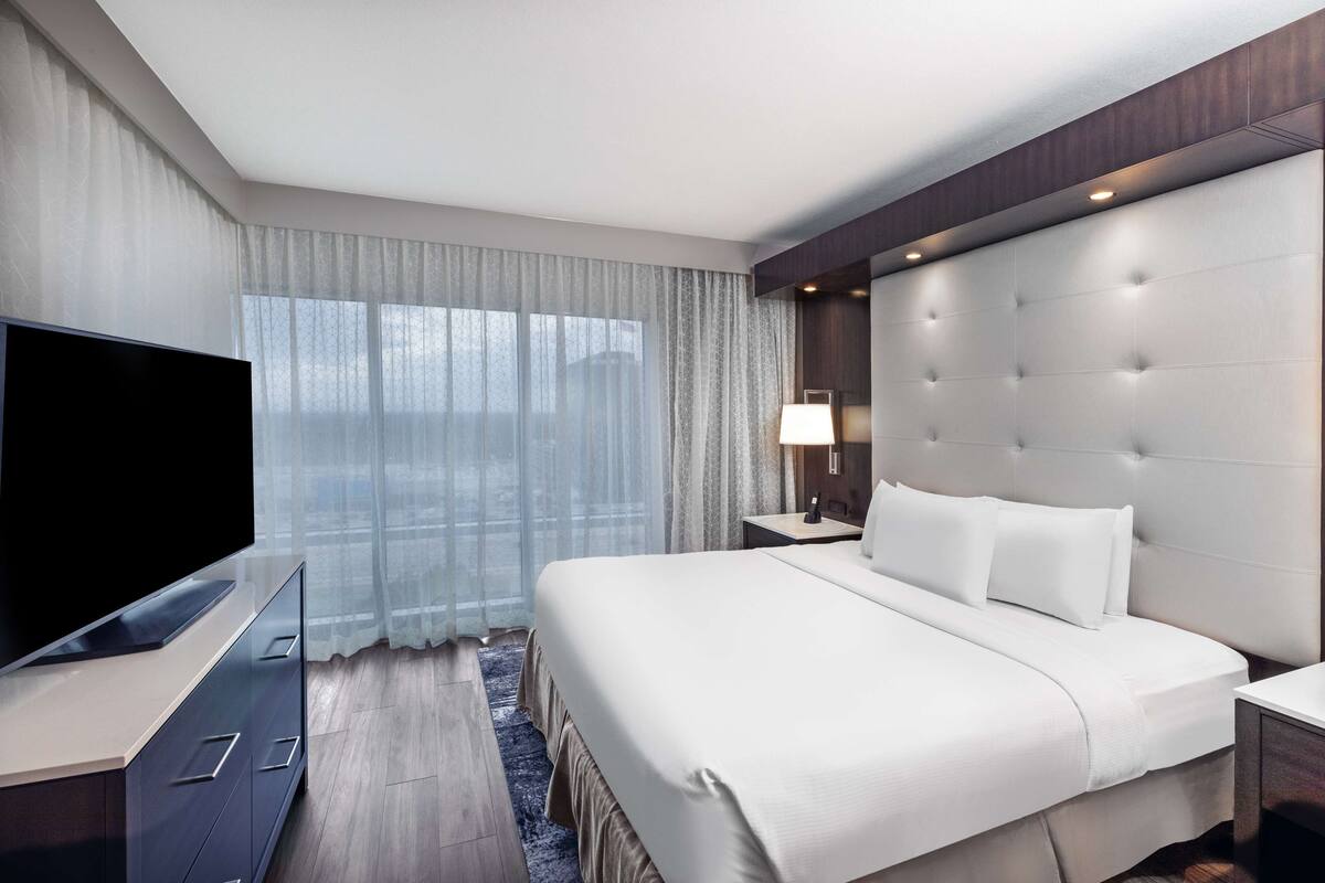 A modern hotel room with a large bed, a flat-screen TV, a bedside table, and a window with sheer curtains.