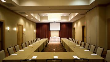 Un salón de conferencias con una larga mesa, sillas, una pantalla de proyector y una zona de escenario.