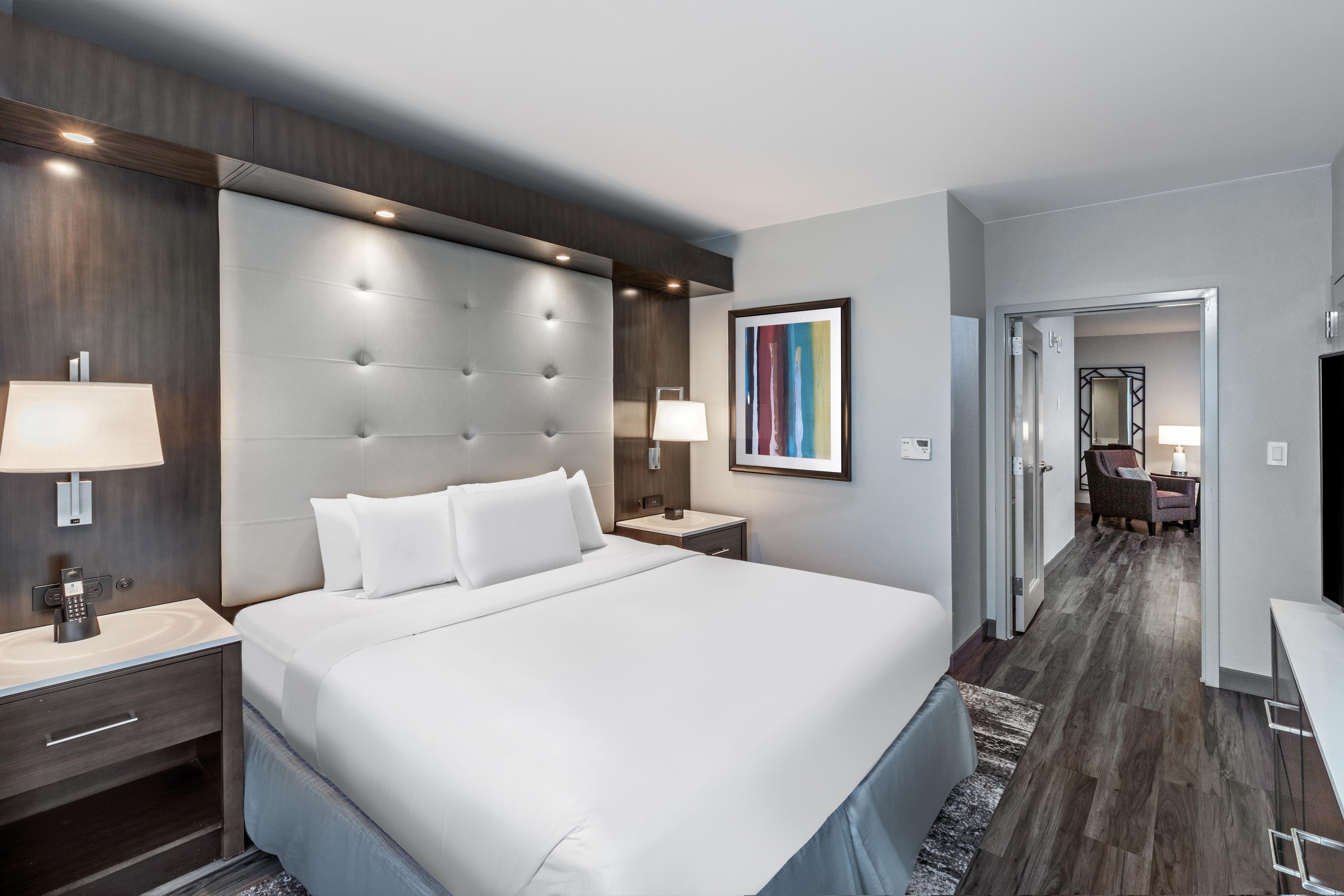 Un cuarto de hotel con una cama grande, lámparas de noche, una mesita de noche, una obra de arte enmarcada y vista al pasillo.