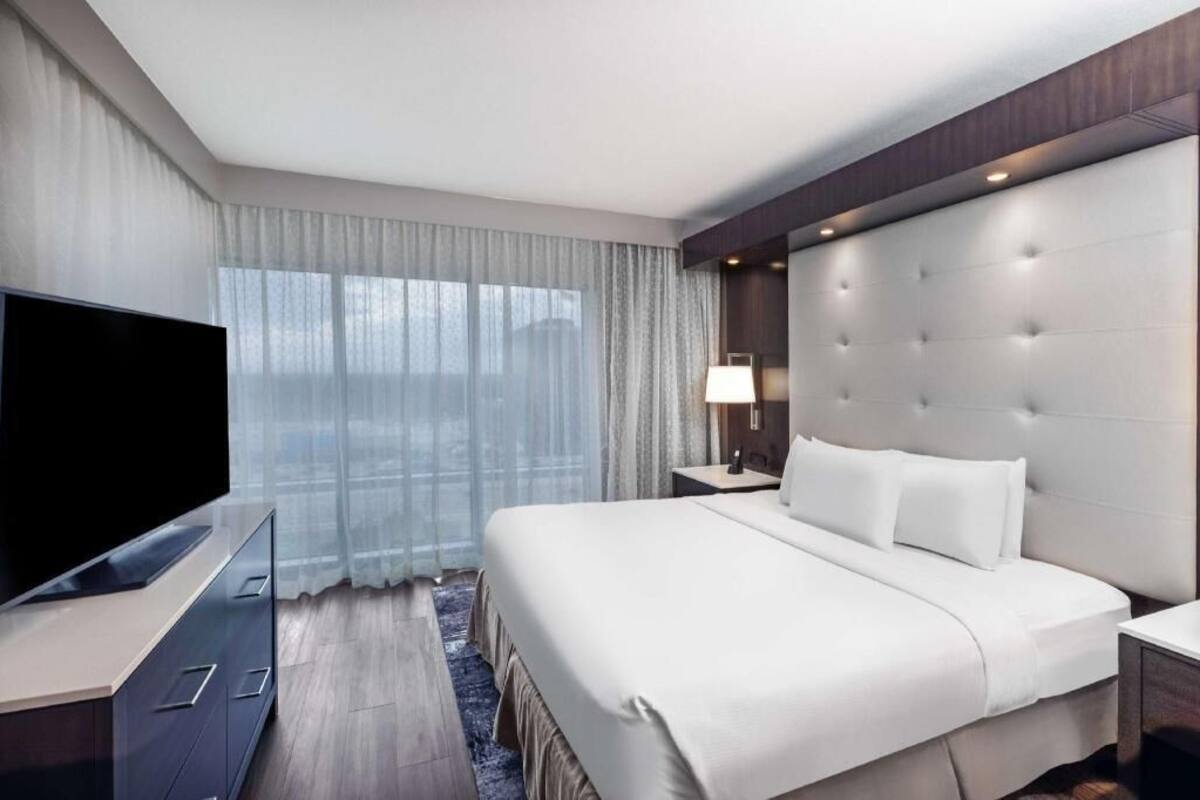 A modern hotel room with a large bed, a flat-screen TV, a bedside table, and a window with sheer curtains.