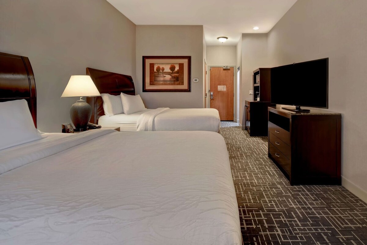 Ein Hotelzimmer mit zwei Betten, einem Schreibtisch mit Lampe, einem Fernseher und einem gerahmten Bild an der Wand.