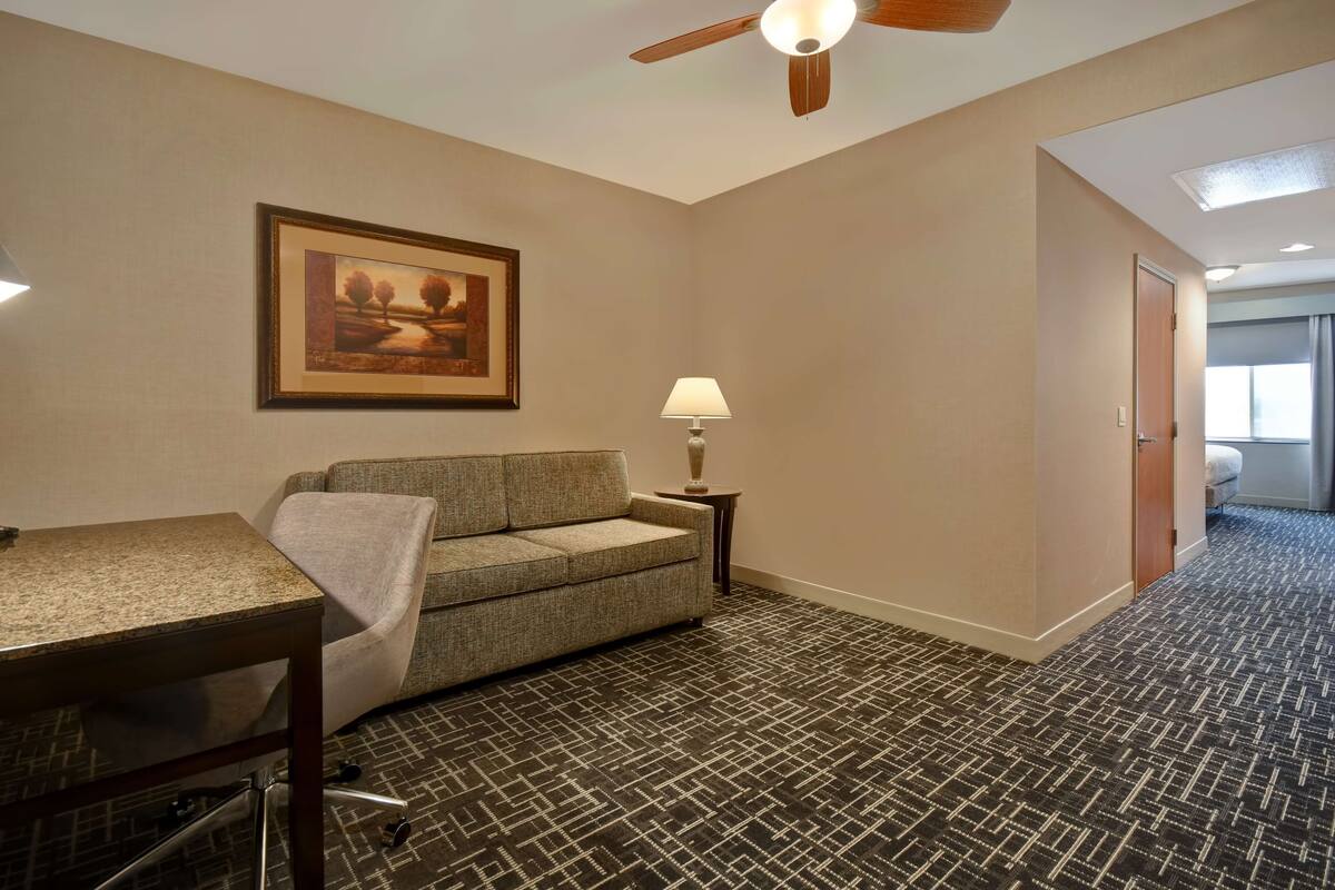 A hotel room with a sofa, a small table, a lamp, a ceiling fan, and a framed picture on the wall.