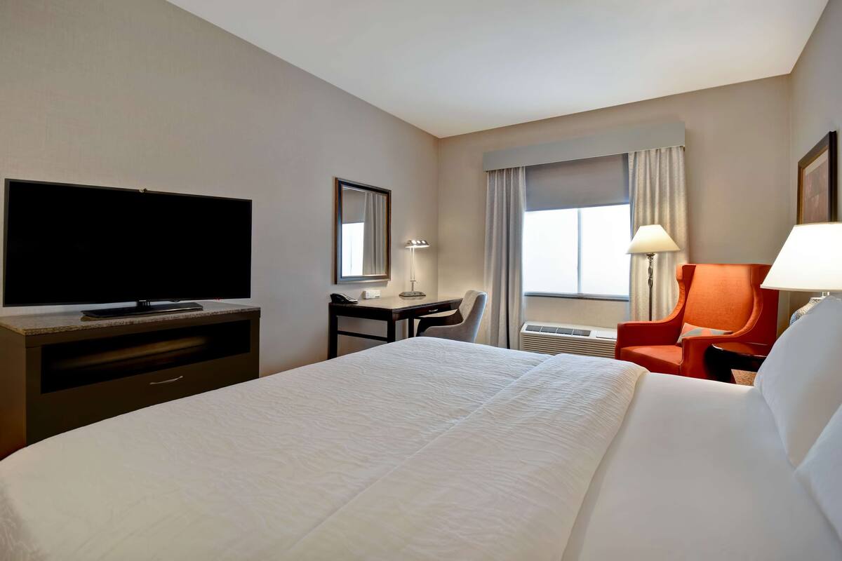 A hotel room with a large bed, a desk with a chair, a television, a lamp, and a window with curtains.