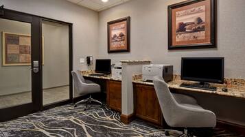 A hotel room with two desks, each with a computer, a printer, and a telephone. There are two chairs and a carpeted floor.